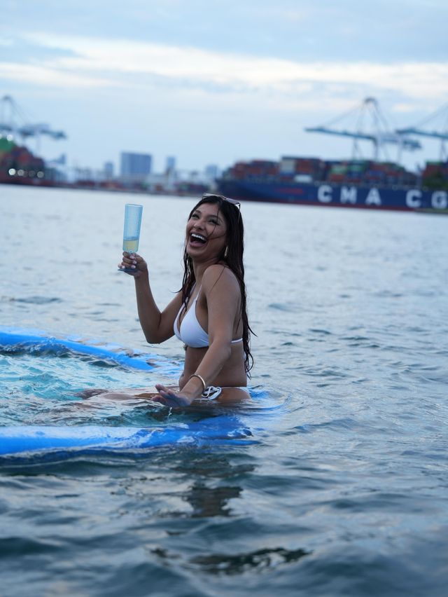 Laughing woman in a white bikini holding a champagne flute while sitting on a blue paddleboard in a busy harbor with container ships and port cranes on the skyline.