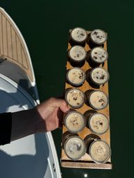 Hand holding a wooden flight paddle with twelve espresso shots above deep green marina water beside a boat deck.