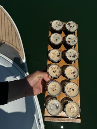 Hand holding a wooden flight paddle with twelve espresso shots above deep green marina water beside a boat deck.