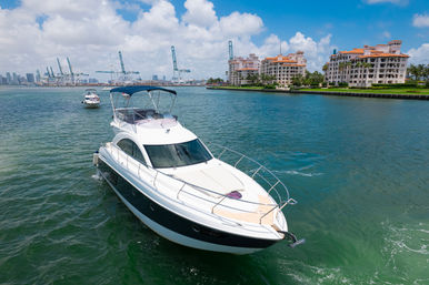Luxury white yacht cruising turquoise coastal waters past waterfront condos and a distant city skyline with shipping cranes under a sunny blue sky