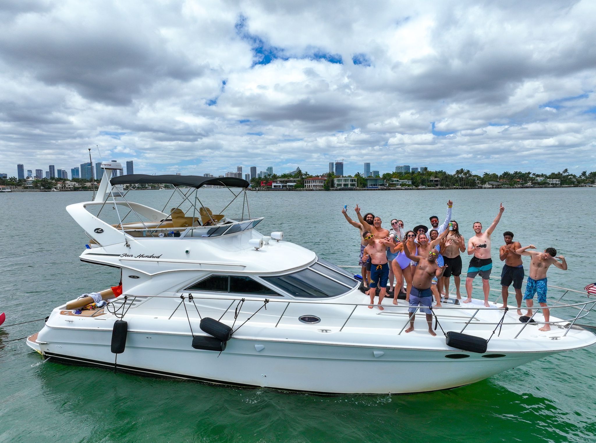 Cheering group of people partying on a white motor yacht in green water, waving and holding drinks with a tropical coastal skyline of palm trees and high-rise buildings under a partly cloudy sky.
