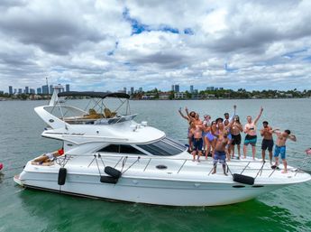 Cheering group of people partying on a white motor yacht in green water, waving and holding drinks with a tropical coastal skyline of palm trees and high-rise buildings under a partly cloudy sky.