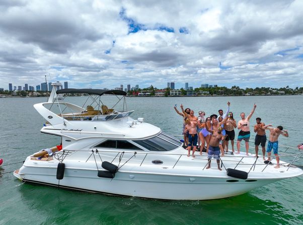 Cheering group of people partying on a white motor yacht in green water, waving and holding drinks with a tropical coastal skyline of palm trees and high-rise buildings under a partly cloudy sky.