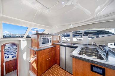 Luxury motor yacht galley with stainless-steel sink, microwave, granite countertops and warm wood cabinetry, large windows revealing a marina and blue sky outside