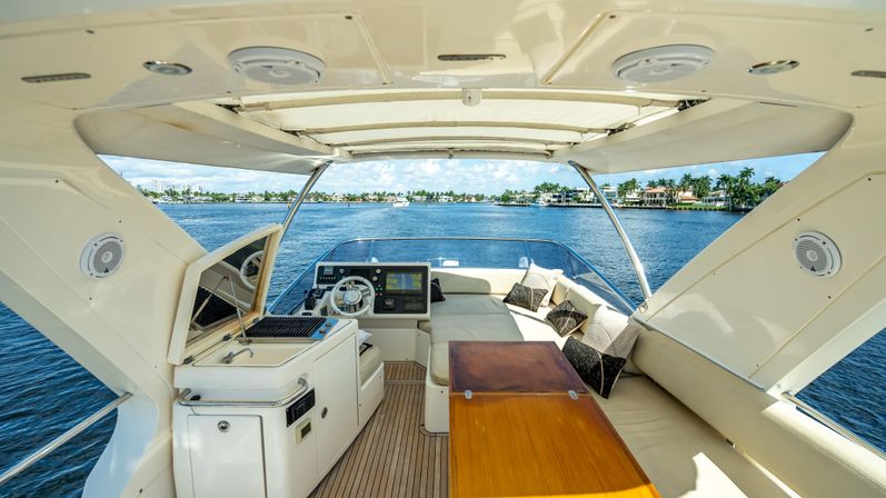 Sun-drenched luxury yacht cockpit with teak deck, helm, built-in grill and cushioned seating around a wooden table, overlooking calm blue water and waterfront homes with palm trees.