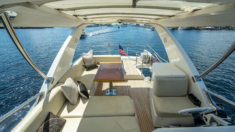 Relaxing aft deck of a luxury motor yacht with teak flooring, cushioned lounge seating and wooden table under a sun canopy, American flag at the stern and blue marina waters beyond.