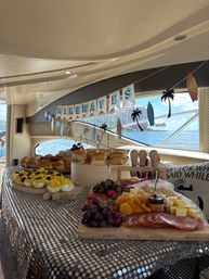 Yacht brunch spread with charcuterie board, cheeses, fruit, deviled eggs and sliders under a "Make Waves" banner, ocean view