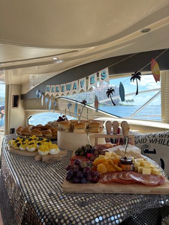 Yacht brunch spread with charcuterie board, cheeses, fruit, deviled eggs and sliders under a "Make Waves" banner, ocean view