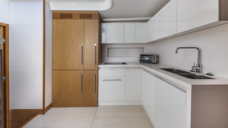 Compact modern galley kitchen with white gloss cabinets, wood-paneled built-in refrigerator, stainless sink and faucet, toaster oven on the counter, and light wood flooring