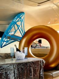 Luxury yacht interior with a large gold inflatable ring and blue shark-tail float, champagne bottle in an ice bucket and two blue flutes on a sequined tablecloth — playful onboard party scene