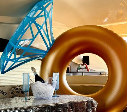 Luxury yacht interior with a large gold inflatable ring and blue shark-tail float, champagne bottle in an ice bucket and two blue flutes on a sequined tablecloth — playful onboard party scene