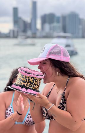 Woman in a pink mesh cap and leopard-print bikini playfully biting a leopard-print birthday cake with pink and black frosting on a boat, with a blurred city skyline and yachts on the waterfront in the background.