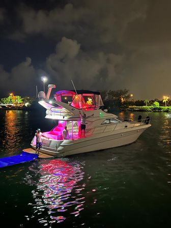 Luxury white yacht at a nighttime marina with pink and purple LED party lights, people on deck, and vibrant reflections on the water beneath a cloudy sky.