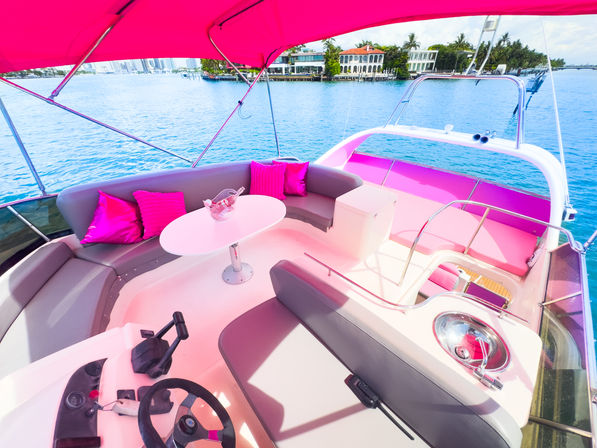 Vibrant pink-accented yacht lounge with cushions and table, sunlit canopy, overlooking turquoise bay and waterfront mansions with a distant city skyline