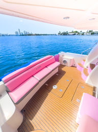 Cheerful pink-cushioned yacht deck with teak-style flooring overlooking vivid blue water and a distant coastal city skyline on a sunny day.