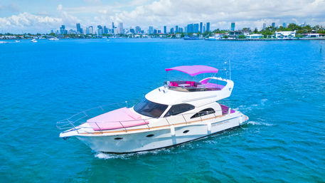 White motor yacht with pink canopy and cushions cruising turquoise bay with Miami skyline and waterfront homes in the background on a sunny day
