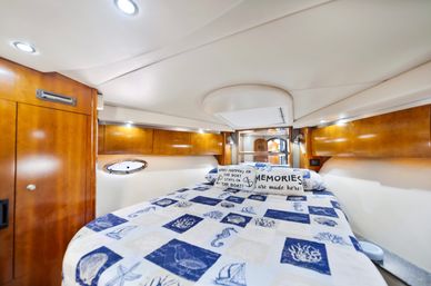 Cozy nautical-themed yacht stateroom with blue-and-white seashell quilt, decorative pillows reading 'Memories are made here', warm wood cabinetry, recessed lighting and a small porthole window.