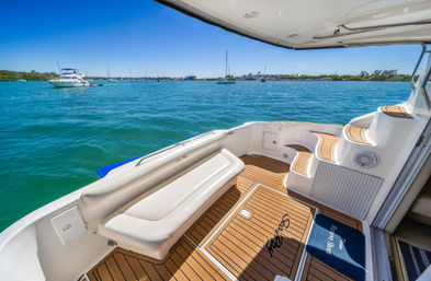 Sunny turquoise harbor view from a yacht's teak deck with cushioned bench, curved steps, and anchored sailboats in the distance.