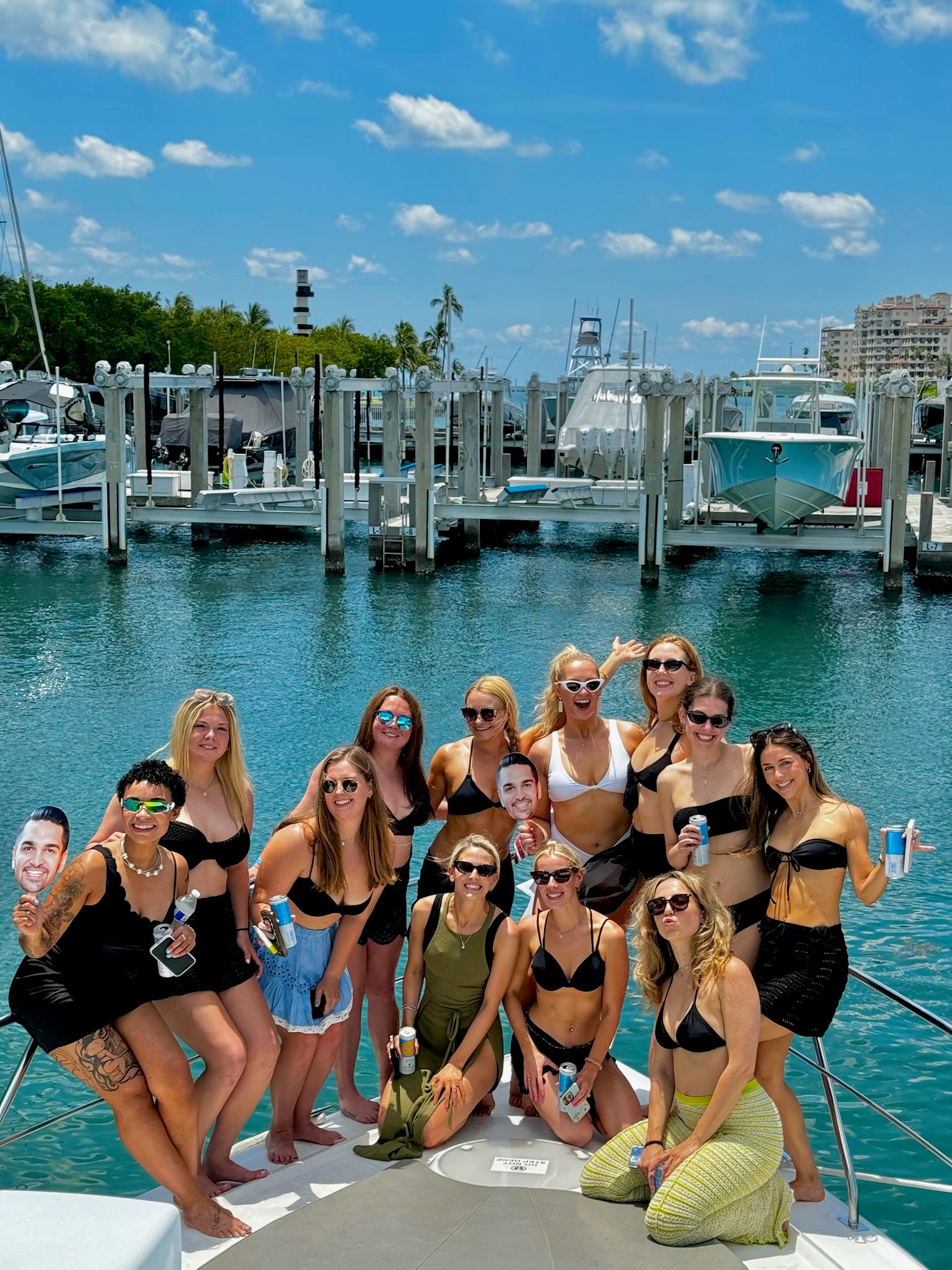 Sunny yacht party: a smiling group of women in swimsuits posing on a boat bow at a tropical marina, turquoise water, docked yachts and palm trees in the background, many holding drinks and playful cardboard face cutouts.