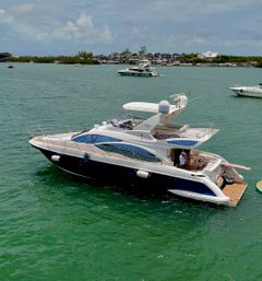 Luxury white-and-navy motor yacht anchored in turquoise coastal bay, open aft deck with passengers, other boats and a marina visible in the background