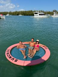 Three adults relaxing on a pink circular 'SUNCHILL' floating lounge in turquoise coastal bay, with anchored yachts and a tree-lined shoreline under a sunny blue sky.