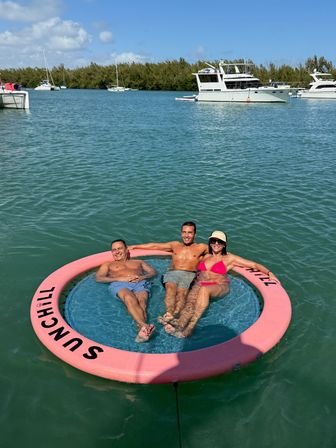 Three adults relaxing on a pink circular 'SUNCHILL' floating lounge in turquoise coastal bay, with anchored yachts and a tree-lined shoreline under a sunny blue sky.