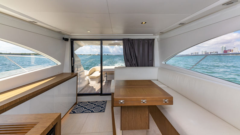 Sunlit luxury yacht interior with white L-shaped seating, wooden foldable dining table, panoramic windows framing turquoise ocean and distant city skyline through sliding glass door to the aft deck.