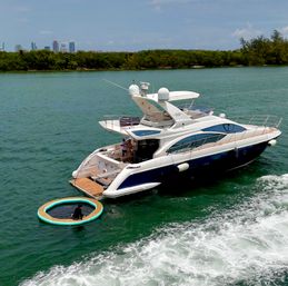 Luxury white-and-blue motor yacht anchored in turquoise coastal waters near a tree-lined shore and distant city skyline, an inflatable circular floating lounge beside the stern with a person relaxing aboard on a sunny day.
