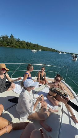 Group of friends relaxing on the bow of a white yacht on a sunny day, passing around shots; turquoise coastal inlet with tree-lined shoreline and several motorboats under a clear blue sky.