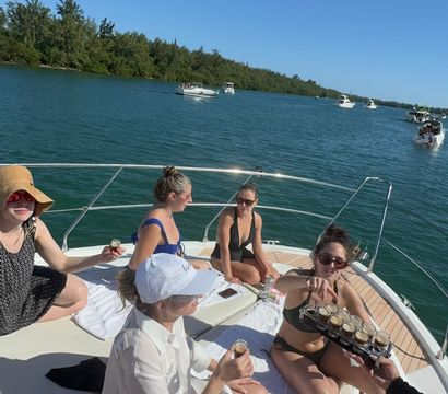 Group of friends relaxing on the bow of a white yacht on a sunny day, passing around shots; turquoise coastal inlet with tree-lined shoreline and several motorboats under a clear blue sky.