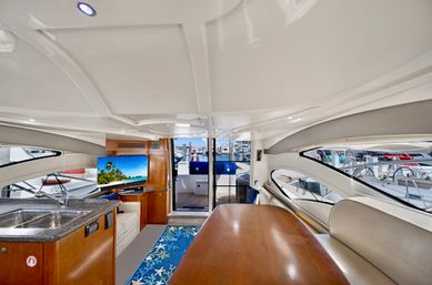 Sunlit luxury yacht salon with glossy white ceiling, teak dining table, beige leather seating, compact galley and sink, flatscreen TV, and sliding glass doors opening to a docked marina view.