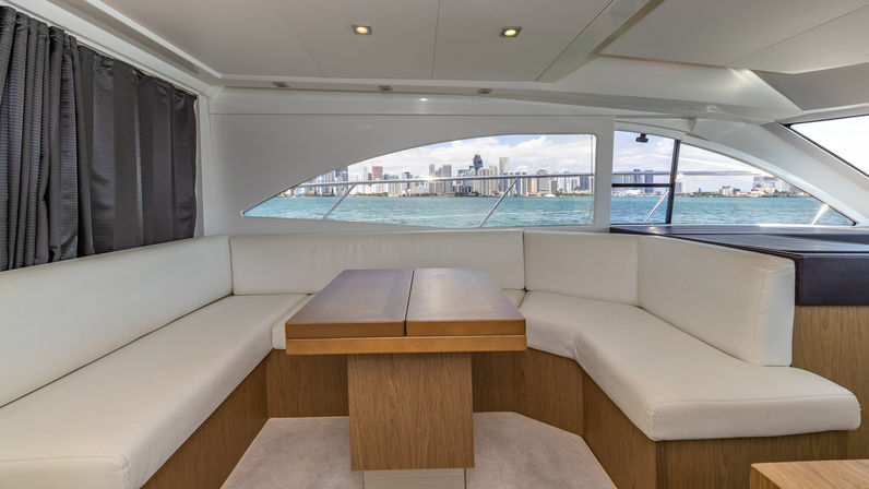 Luxury yacht salon with white leather L-shaped seating and wooden table, large windows framing a turquoise bay and distant city skyline under a blue sky.