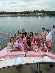 Cheerful group of friends on a pink-cushioned yacht bow, wearing pink swimwear and shorts, holding drinks and posing by a calm marina under a cloudy sky.