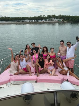 Cheerful group of friends on a pink-cushioned yacht bow, wearing pink swimwear and shorts, holding drinks and posing by a calm marina under a cloudy sky.