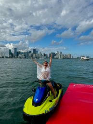 Person in white lace cover-up cheering on a bright yellow and blue jet ski beside a red dock in Biscayne Bay with the Miami skyline and a partly cloudy blue sky in the background.