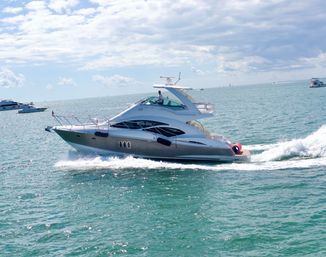 Sleek white luxury motor yacht cruising across turquoise coastal waters, cutting a foamy wake under a sunny, partly cloudy sky.