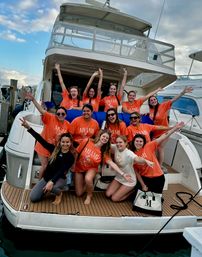 Cheerful group of friends posing on the swim platform of a white yacht at a marina, most wearing matching orange "MIAMI" shirts with arms raised and big smiles under a partly cloudy sky.