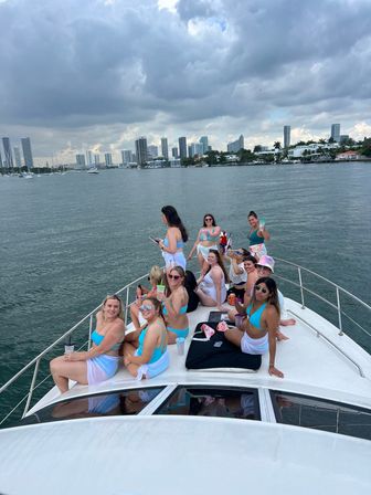 Friends enjoying a daytime yacht party on the bow in matching turquoise swimsuits, holding drinks while cruising a calm bay with a modern city skyline and moody clouds overhead.