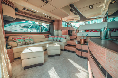Sunlit luxury motor yacht salon with cream leather L-shaped seating, patterned accent pillows, matching ottomans, warm wood-accent cabinetry and a champagne bucket, large windows framing marina views.