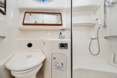 Compact yacht bathroom (marine head) with white toilet, built-in sink and mirrored cabinet, handheld shower, grab bars and round porthole.