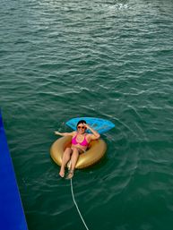 Aerial view of a smiling person in orange sunglasses and a pink bikini relaxing on a gold inflatable tube with a blue float, tethered and drifting in green open water on a sunny day.