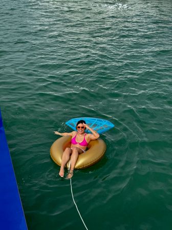Aerial view of a smiling person in orange sunglasses and a pink bikini relaxing on a gold inflatable tube with a blue float, tethered and drifting in green open water on a sunny day.