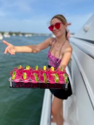 Person on a boat offering a tray of bright pink shot glasses garnished with lime wedges, sunny harbor and boats in the blurred background — summer boat party vibe.