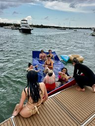 Boat party in a coastal bay — group of women in swimsuits on a blue inflatable mat beside a yacht, anchored boats and cloudy sky in the background.