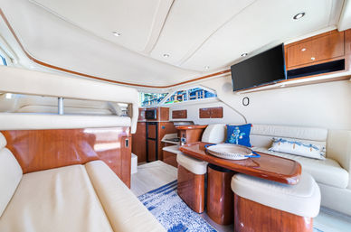Bright luxury yacht cabin with cream leather seating, glossy cherry wood table and cabinetry, flat-screen TV, nautical pillows and a marina view through the windows.