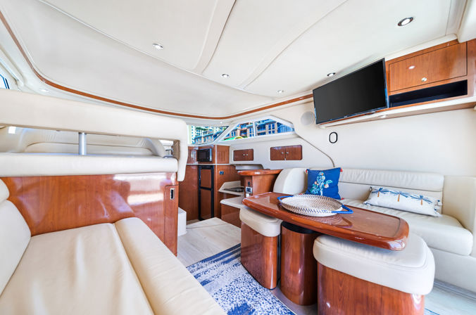 Bright luxury yacht cabin with cream leather seating, glossy cherry wood table and cabinetry, flat-screen TV, nautical pillows and a marina view through the windows.