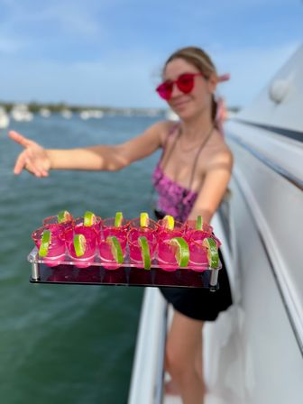 Tray of vibrant pink shot glasses garnished with lime wedges held aboard a boat, blurred marina and water in the background for a sunny summer party vibe