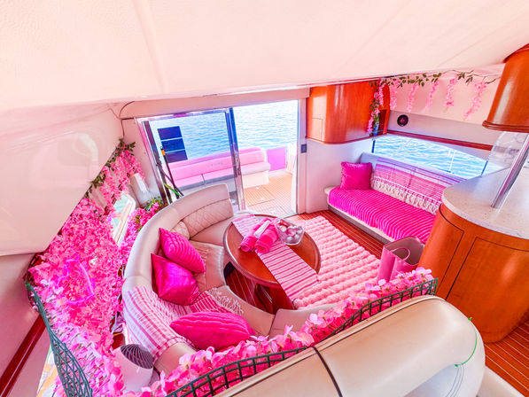 Bright pink-themed yacht interior salon with curved beige leather seating, striped cushions and rugs, pink floral garlands, wooden table and sliding glass door opening to sunlit deck and blue ocean