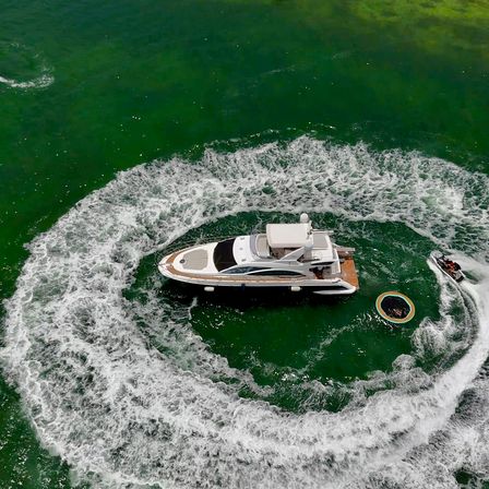 Drone aerial view of a white luxury yacht in emerald coastal water, a jet ski carving a dramatic circular wake around it and a colorful inflatable tow ring floating nearby