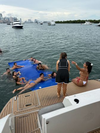 People relaxing on a large blue floating mat beside a boat swim platform, anchored yachts nearby and a coastal city skyline across the bay.